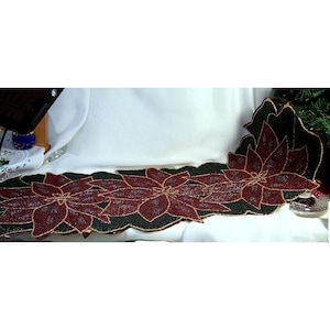 May include: A long, rectangular table runner featuring a repeating pattern of three large, dark red poinsettia flowers with gold-toned outlines. The background of the runner is composed of black beads, and the petals are filled with red beads.