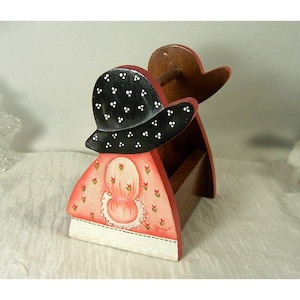 May include: A wooden folk art organizer shaped like a woman in a bonnet. The bonnet is black with white polka dots. The dress is pink with floral accents and a white lace collar. The organizer has a small shelf and a wooden spool holder.