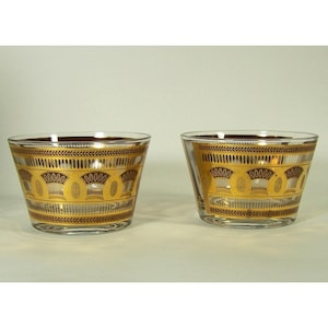 May include: Two clear glass bowls with a gold and brown decorative pattern. The bowls have a wide, cylindrical shape with a slightly flared rim. The gold design features repeating geometric shapes and a border of leaves.