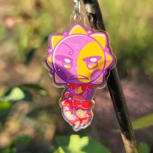 Può includere: Un ciondolo in acrilico trasparente con un personaggio dei cartoni animati con un design a volto diviso, metà viola e metà giallo. Il personaggio ha un contorno a forma di sole e indossa un abito rosso e bianco. Un fermaglio color argento è attaccato in alto.