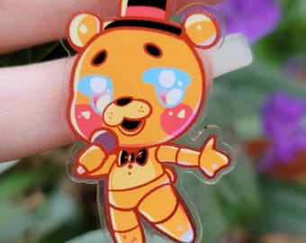 May include: A clear acrylic keychain featuring a cartoon bear character with orange fur, a black top hat, and a bow tie. The bear has pink cheeks, blue tears, and is holding a microphone. The keychain has a silver metal ring.