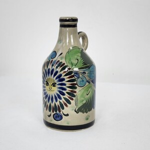 May include: A small ceramic jug with a handle, painted with a white background and colourful floral designs. The jug features a sun face with a yellow face and blue eyes.