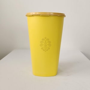 Puede incluir: Un recipiente alto de plástico amarillo con tapa marrón claro. El recipiente tiene un diseño floral decorativo en el centro. El recipiente mide aproximadamente 25 cm de alto.