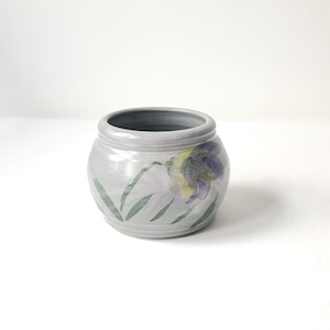 May include: A small, round, gray ceramic pot with a purple and yellow flower design. The pot has a glazed finish and is perfect for small plants or succulents.