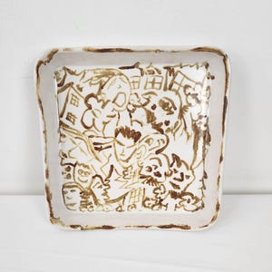 Puede incluir: Un plato cuadrado de cerámica blanca con un glaseado marrón. El plato está decorado con un diseño caprichoso dibujado a mano de caras y figuras.