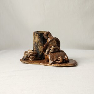 Peut inclure: Une figurine de gnome en céramique avec un chapeau et une barbe bruns, assise sur une base brune avec un cylindre en céramique brun. Le gnome tient un petit objet brun dans ses mains.