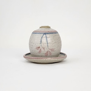 May include: A white ceramic vase with a pink and blue floral design and a matching saucer. The vase has a speckled glaze and a round shape.