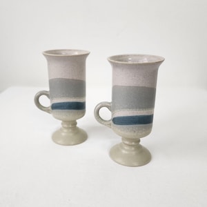 Puede incluir: Dos tazas de café de cerámica con base de pedestal y asa. Las tazas tienen una combinación de colores gris claro y azul con rayas horizontales. Las tazas miden aproximadamente 15 cm de alto.