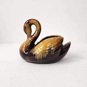 May include: A brown and yellow glazed ceramic swan planter. The swan has a long neck and a wide body. The glaze is a mottled brown and yellow colour.