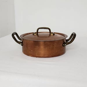 Peut inclure: Une marmite ronde en cuivre avec son couvercle assorti, dotée de deux poignées latérales et d'une poignée sur le couvercle. La marmite présente une finition métallique polie avec des marques d'usage et une teinte brun-rouge chaleureuse.
