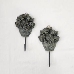 May include: A pair of decorative wall hooks, each with a grey pot filled with sculpted flowers. The hooks have a distressed finish, with a dark metal hook extending downwards. They are mounted on a white wall.