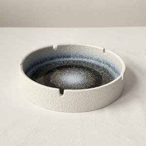 May include: A white ceramic ashtray with a blue and black speckled glaze. The ashtray has a round shape with a slightly raised rim and four small indentations for cigarettes.