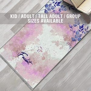 May include: A rectangular prayer rug with a watercolor design in shades of pink, purple, and white. The rug features a floral pattern in blue and the word "Faidat" in a script font. Text at the top reads "KID / ADULT / TALL ADULT / GROUP SIZES AVAILABLE."