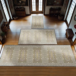 May include: A set of three beige prayer rugs with intricate floral patterns and arched designs. The rugs are arranged in a staircase pattern on a wooden floor.