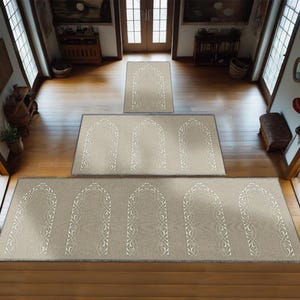May include: A set of three beige prayer rugs with a white intricate design of arches and vines. The rugs are arranged in a staircase pattern on a wooden floor.