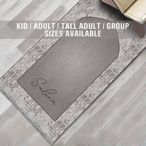 May include: A gray prayer rug with a white floral pattern border. The rug has a personalized name, "Salina", in cursive script. The text "KID / ADULT / TALL ADULT / GROUP SIZES AVAILABLE" is displayed above the rug.