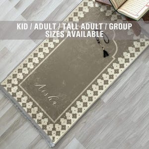 May include: A beige prayer rug with a white geometric pattern border. The rug has a personalized name, "Aisha", in cursive script. The rug is on a light wood floor.