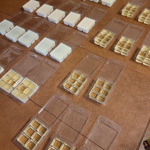 May include: An overhead shot displays multiple wax melt clamshells. Some contain rectangular bars of off-white wax, while others hold golden-brown wax cubes. The clamshells are arranged on a brown surface, suggesting a collection of scented wax melts.
