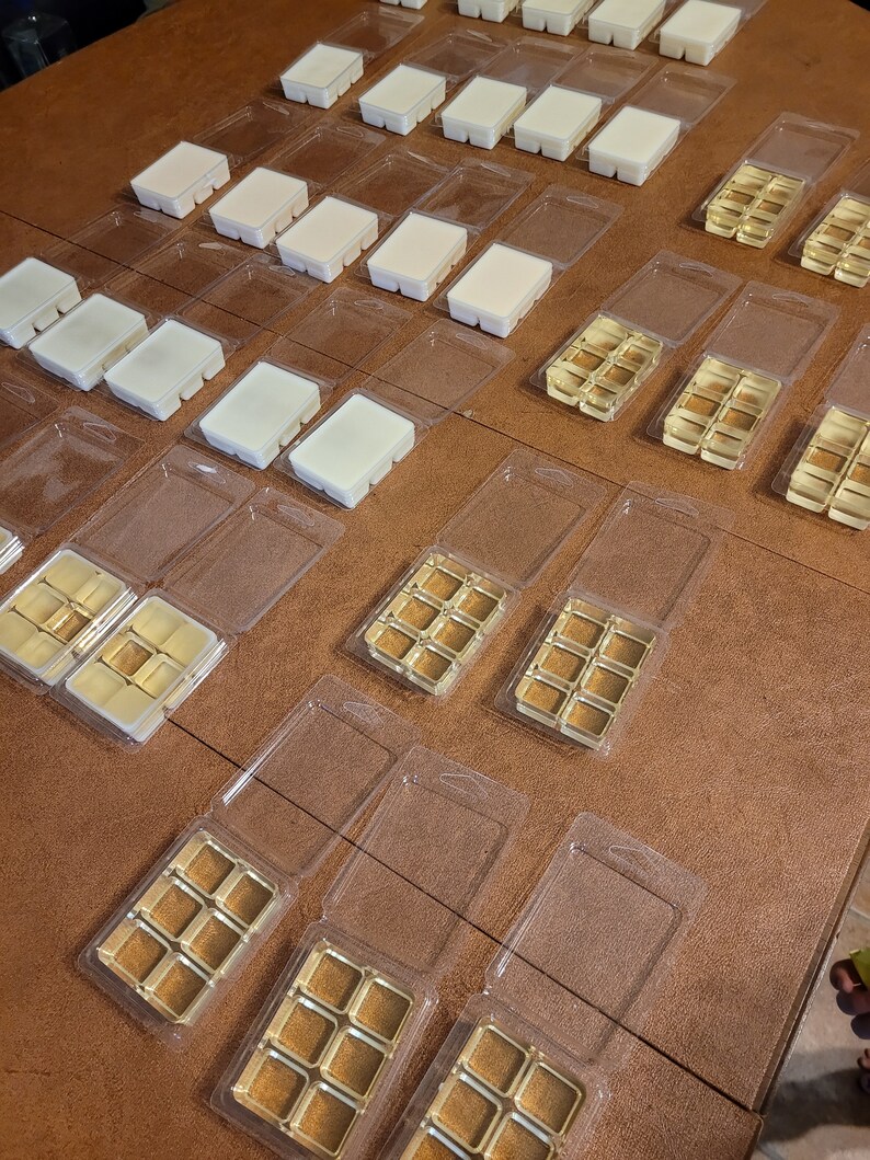 May include: An overhead shot displays multiple wax melt clamshells. Some contain rectangular bars of off-white wax, while others hold golden-brown wax cubes. The clamshells are arranged on a brown surface, suggesting a collection of scented wax melts.