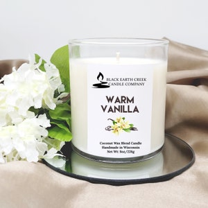 Pu&ograve; includere: Una candela bianca in un barattolo di vetro trasparente con un'etichetta che recita "Black Earth Creek Candle Company" e "Warm Vanilla". La candela &egrave; su un vassoio argentato e circondata da fiori bianchi.