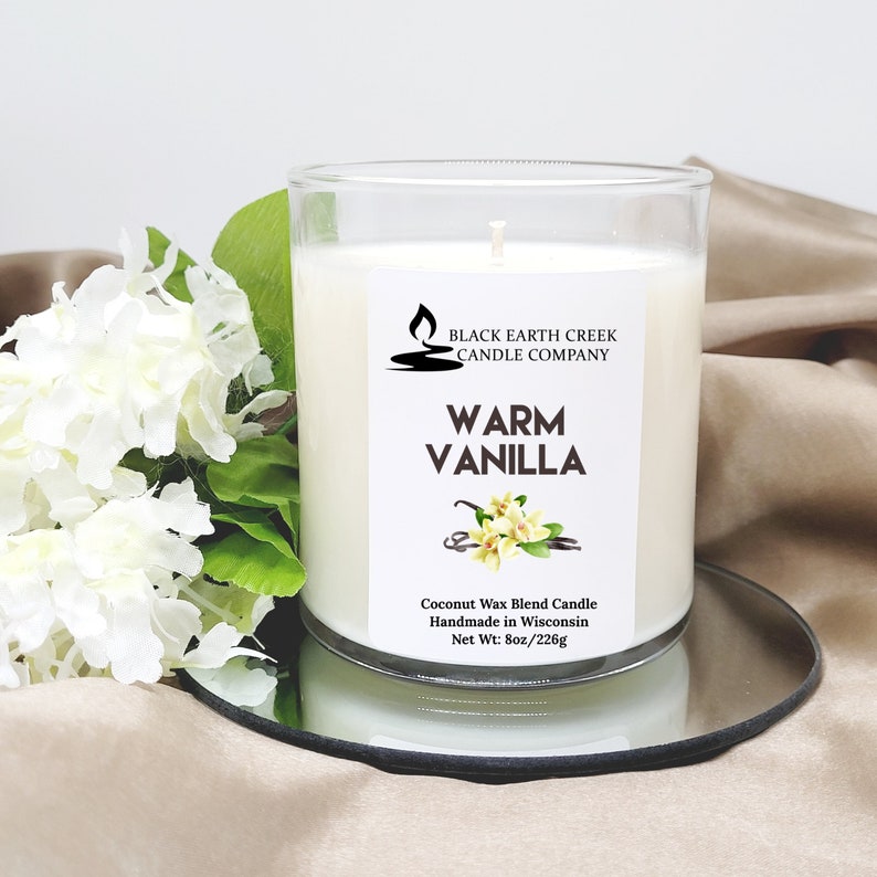 Pu&ograve; includere: Una candela bianca in un barattolo di vetro trasparente con un'etichetta che recita "Black Earth Creek Candle Company" e "Warm Vanilla". La candela &egrave; su un vassoio argentato e circondata da fiori bianchi.