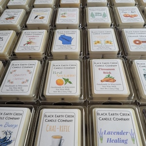 May include: An overhead shot of multiple soy wax melts in clear plastic containers. Each melt has a label with a unique scent name and graphic, such as "Cranberry Spice," "Basil Nectarine," and "Lavender Healing." The net weight is 2.5 oz / 70g.