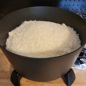May include: A black pot filled with off-white, granular wax. The pot has a wide opening and three black legs. The wax appears to be in the process of being melted for candle making.