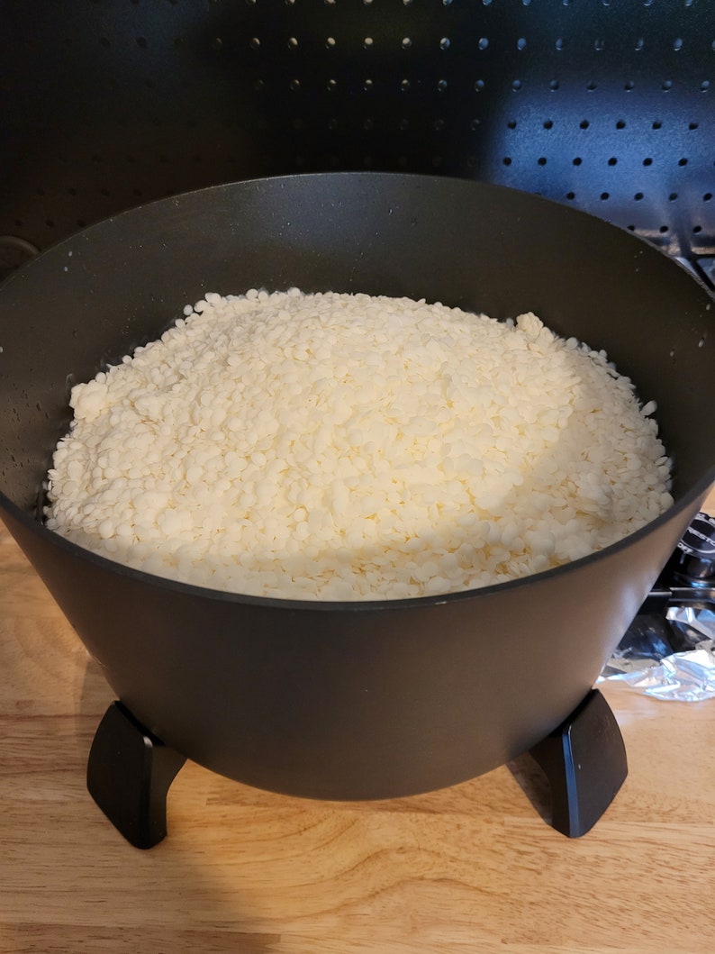 May include: A black pot filled with off-white, granular wax. The pot has a wide opening and three black legs. The wax appears to be in the process of being melted for candle making.