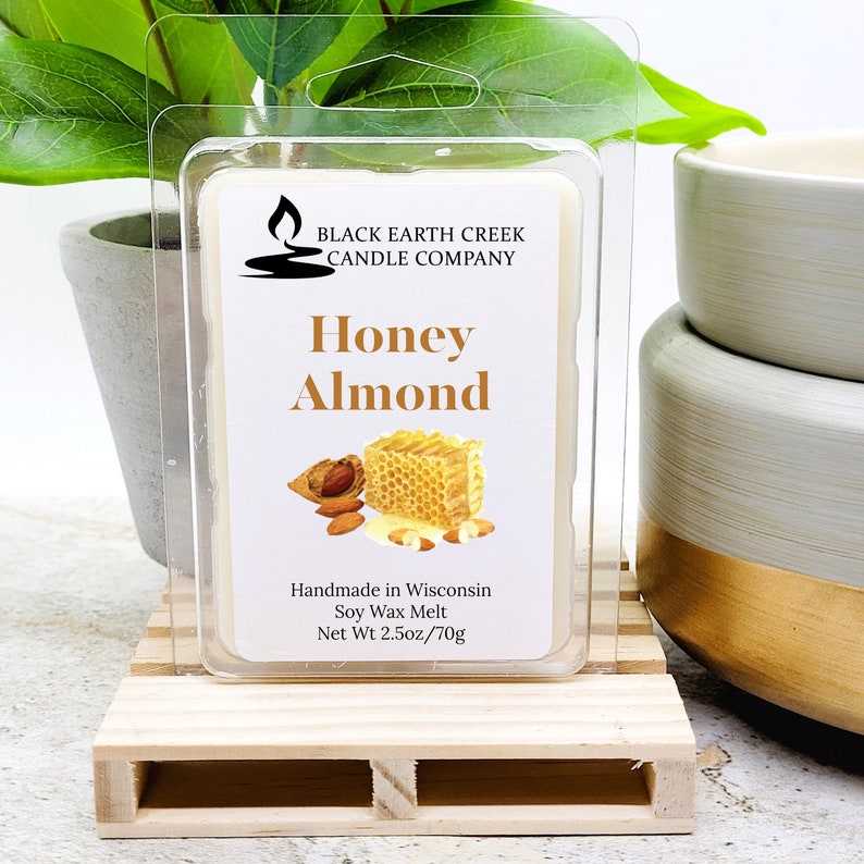 May include: A clear plastic package containing a soy wax melt. The label reads "Honey Almond" with images of almonds and honeycomb. The package is on a small wooden pallet. The text "Handmade in Wisconsin" is also visible.
