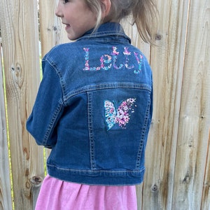 Può includere: Una giacca di jeans blu con il nome "Letty" ricamato sul retro con fiori colorati. Anche una farfalla con accenti floreali è ricamata sul retro.