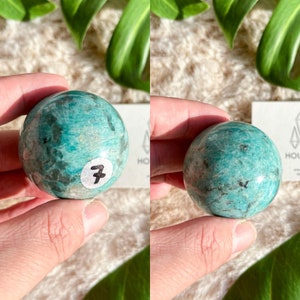 May include: A green and white Amazonite sphere with a number "7" sticker on it. The sphere is being held in a hand.