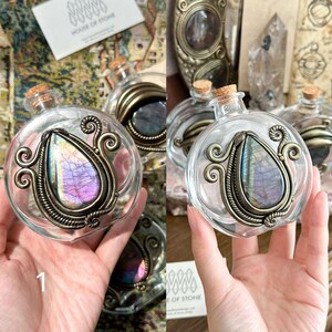 May include: Clear glass bottles with cork stoppers and ornate metal frames. Each bottle features a teardrop-shaped stone with purple, blue, and green hues. The bottles are labeled "HOUSE OF STONE".