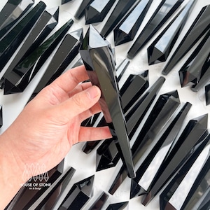May include: A collection of black obsidian crystal points, also known as black glass, arranged on a white surface. One point is being held by a hand. The points are all sharp and have a smooth, polished finish.