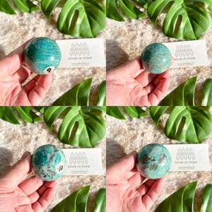 May include: Three green amazonite sphere crystal balls, each with a different pattern of white and gray markings. The spheres are approximately 2 inches in diameter. The spheres are held in a hand, with a business card from "House of Stone" visible in the background.