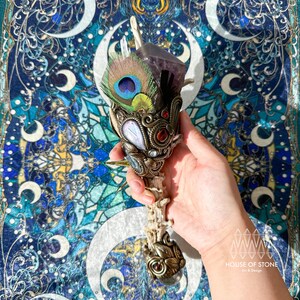 Peut inclure: Une baguette artisanale ornée, comprenant un cristal d'améthyste violet, une plume de paon et diverses pierres précieuses. La baguette présente un design en métal détaillé et est montée sur un manche en os. Le fond est un tissu à motifs bleu et turquoise.