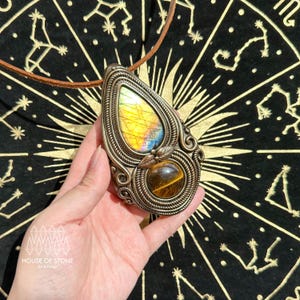 May include: A handcrafted pendant necklace featuring a teardrop-shaped labradorite stone with yellow, blue, and green hues, set in a detailed metal frame. Below is a round tiger's eye stone. The necklace has a brown leather cord. The House of Stone logo is visible.
