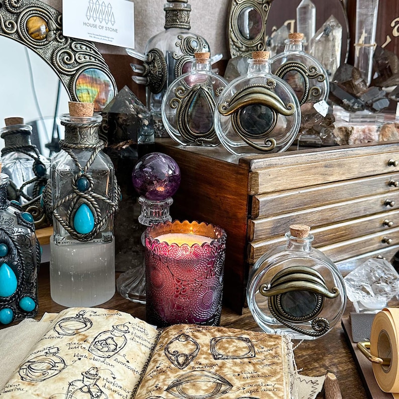 May include: A collection of decorative glass bottles with cork stoppers, adorned with ornate metalwork and gemstones. A lit purple candle and an open book with sketches are also visible, creating a mystical aesthetic.