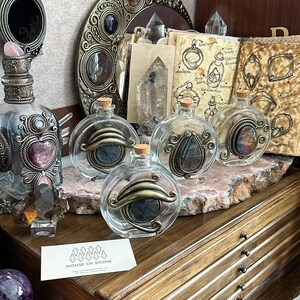 May include: Clear glass bottles with decorative metal accents and cork stoppers. The bottles feature ornate metalwork and contain colorful stones. A business card with the text "HOUSE OF STONE" is visible.