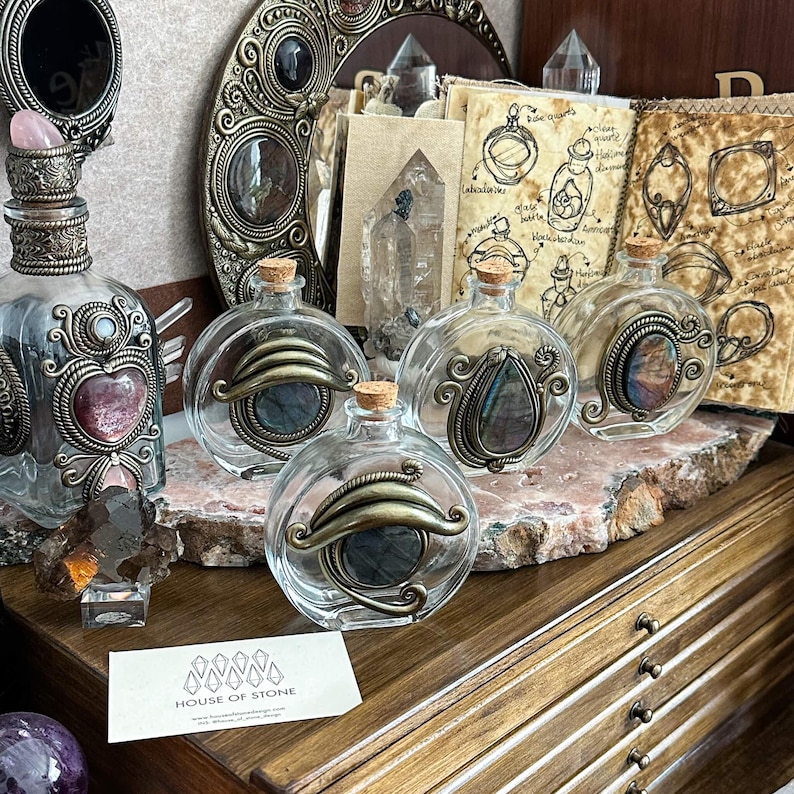 May include: Clear glass bottles with decorative metal accents and cork stoppers. The bottles feature ornate metalwork and contain colorful stones. A business card with the text "HOUSE OF STONE" is visible.