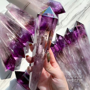 Può includere: Punte di cristallo di ametista di varie dimensioni, che mostrano una sfumatura di tonalità viola. I cristalli sono trasparenti con strutture interne visibili. L'immagine include il testo "HOUSE OF STONE Art & Design".