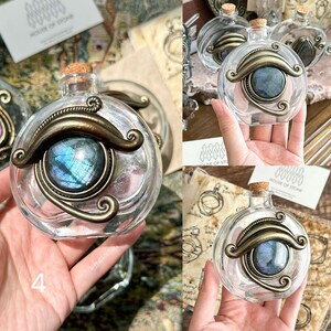 May include: Clear glass bottle with a cork stopper, adorned with a bronze-colored metal eye design. The eye features a blue and green iridescent stone. The bottle is held in a hand, with the number "4" visible.