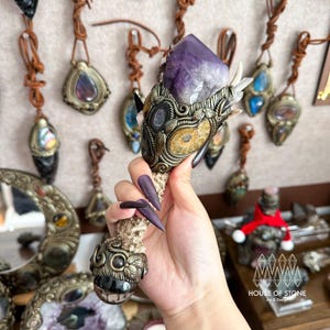 Peut inclure: Une baguette artisanale ornée d'un grand cristal d'améthyste violet facetté. La baguette présente des ornements métalliques avec des détails de fossiles et de feuilles, et un manche en forme d'os. D'autres pendentifs et objets décoratifs sont visibles en arrière-plan.