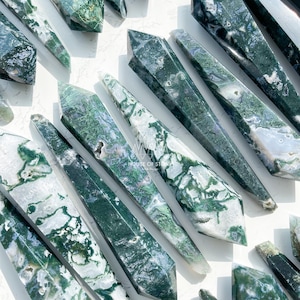 May include: A collection of moss agate crystal towers featuring deep green, translucent, and white mineral inclusions. Each polished stone point displays unique dendritic patterns and natural textures. The text HOUSE OF STONE Art & Design is visible.