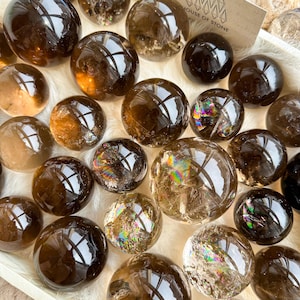May include: A collection of polished, spherical smoky quartz crystals in varying shades of translucent brown and dark charcoal. Several spheres feature internal fractures that display iridescent rainbow light refractions. A card reads HOUSE OF STONE.