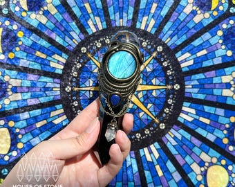 May include: A dark, polished crystal point wrapped in intricate bronze-toned wire. The piece features a round, iridescent blue labradorite cabochon, a small teardrop-shaped lapis lazuli stone, and a dangling clear quartz point on a fine chain.