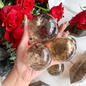 Può includere: Tre sfere di cristallo di quarzo trasparente di dimensioni diverse, con una tonalità marrone fumè. Le sfere sono tenute in una mano, con la sfera più grande al centro.