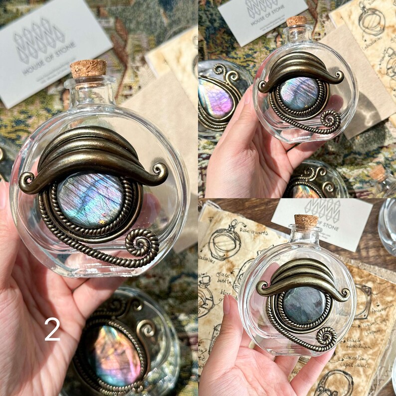 May include: Clear glass bottle with a cork stopper, featuring a decorative metal frame and a shimmering, iridescent stone. The metal frame has an antique bronze finish. The bottle is round and has a unique, artistic design.
