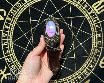 May include: A dark brown crystal point wand topped with an oval labradorite cabochon featuring purple and blue iridescence. The stone is set in an antique bronze metal casing with a small dangling chain and a tiny purple gemstone accent.