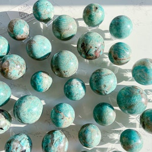 May include: A collection of 20+ green amazonite spheres of varying sizes, each with unique patterns and textures. The spheres are displayed on clear stands on a white surface.