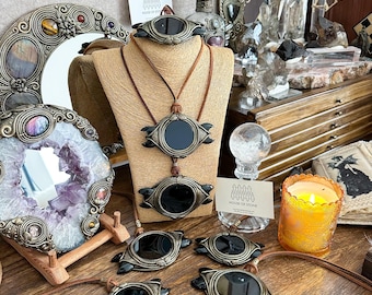 May include: An assortment of handcrafted jewelry pieces, including necklaces and pendants, featuring dark, oval-shaped stones set in ornate, antique-style metal frames. The pieces are displayed on a neutral-toned mannequin and a wooden surface.